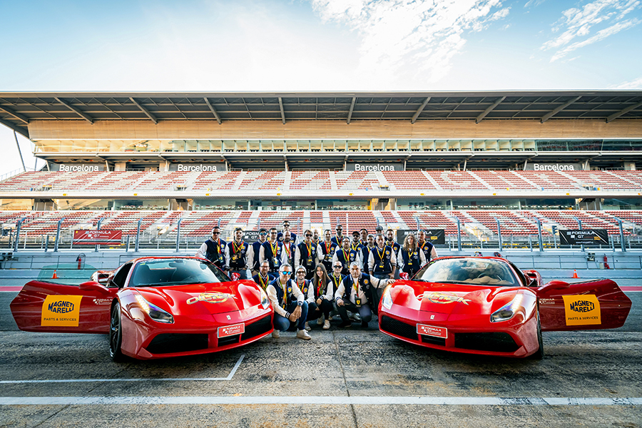 Due Ferrari rosse con il logo Magneti Marelli Parts & Services sulle portiere e il logo Oiltek sul cofano anteriore, sul circuito di Montmelò a Barcellona, con il gruppo dei partecipanti all'evento Oiltek Trophy 2024.
