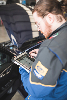 Meccanico esperto con tuta Magneti Marelli Checkstar seduto sul sedile della vettura, impegnato nella diagnosi attraverso un tablet elettronico durante un intervento di autoriparazione, sfruttando le competenze acquisite durante la formazione tecnica.