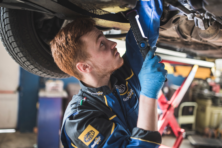 Professionista autoriparatore con tuta Magneti Marelli Checkstar ispeziona attentamente la parte inferiore di un'auto utilizzando una torcia, mentre la vettura è sollevata su un ponte sollevatore, applicando le competenze acquisite durante la formazione tecnica.